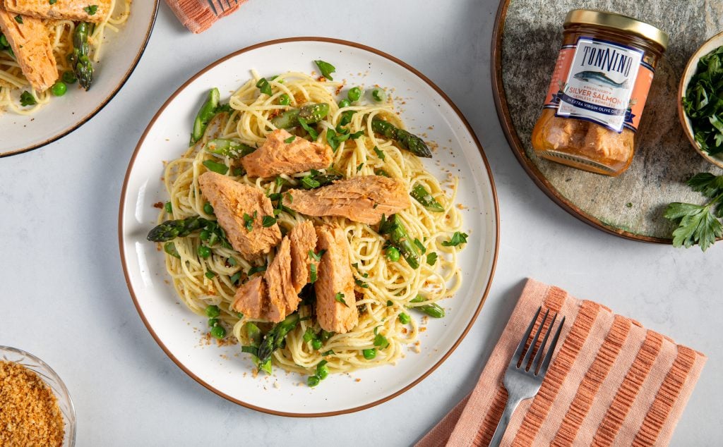 Spaghetti with asparagus, peas, and flaked salmon served on a plate, with a glass jar of salmon placed nearby.